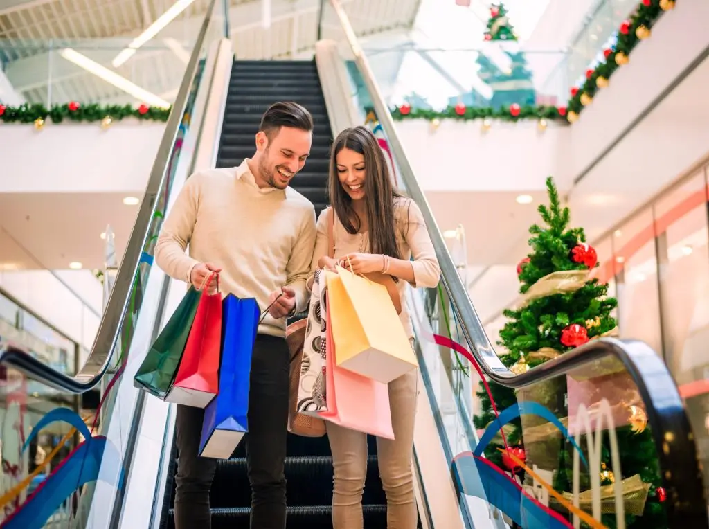 Couple coming down a mall escalator with hands full of shopping bags