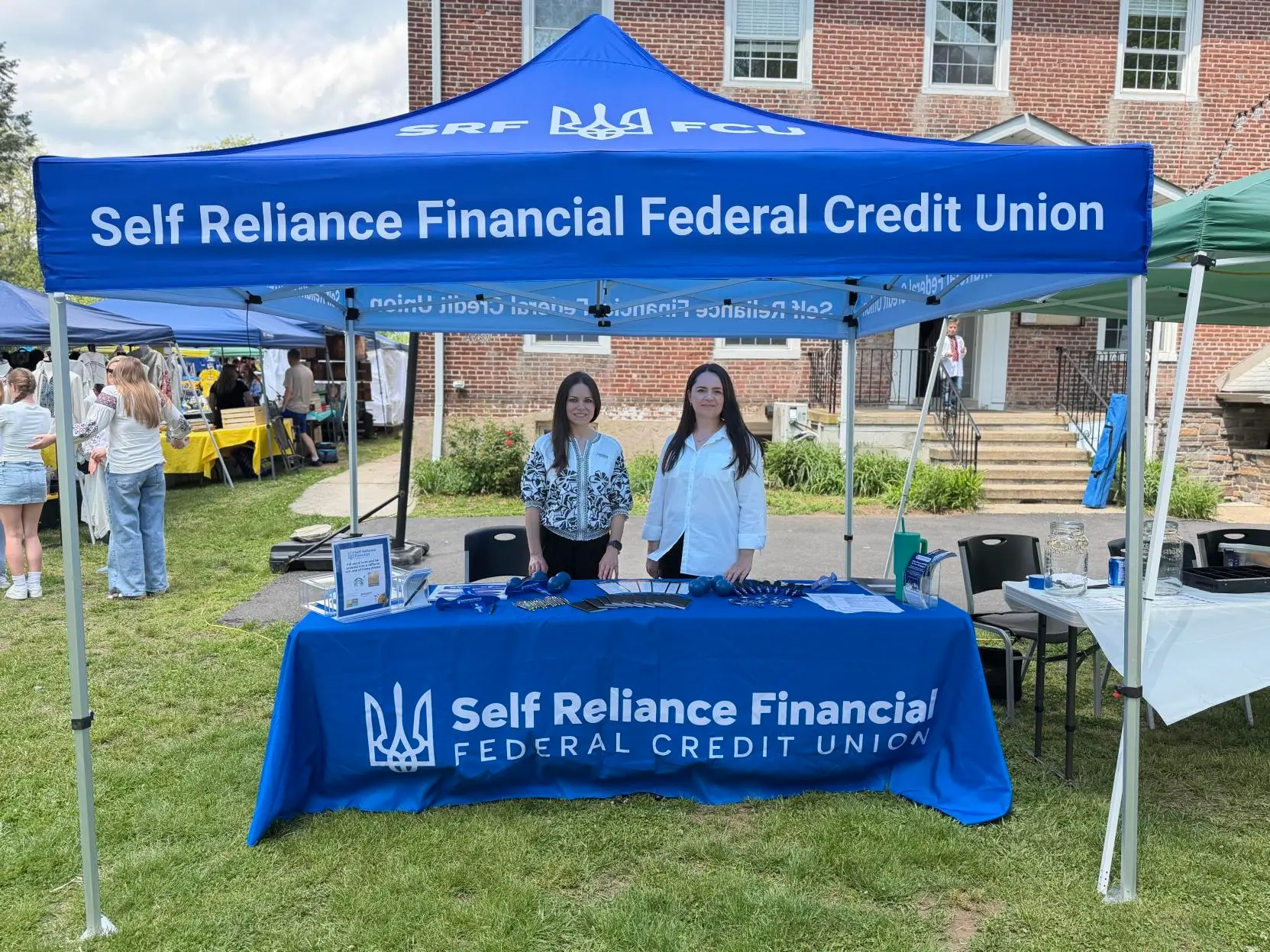 Self Reliance Financial Federal Credit Union team members at an outdoor community event, featuring a blue canopy, branded table, and informational materials.